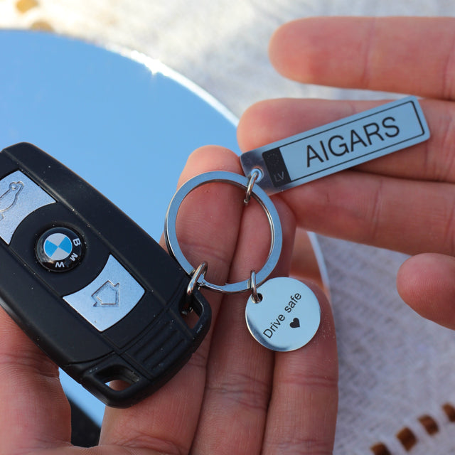 Auto numura zīmes atslēgu piekariņš sudraba krāsā ar Latvijas simboliku. Skats no priekšpuses